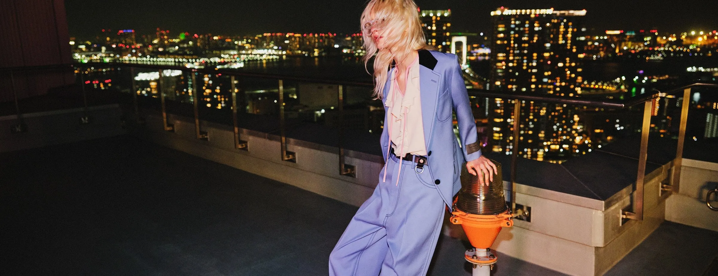 A person with long blonde hair in a pale blue suit and a cream ruffled blouse, leaning against an orange beacon light on a rooftop at night with one hand on top of it, a railing and a lit city skyline behind