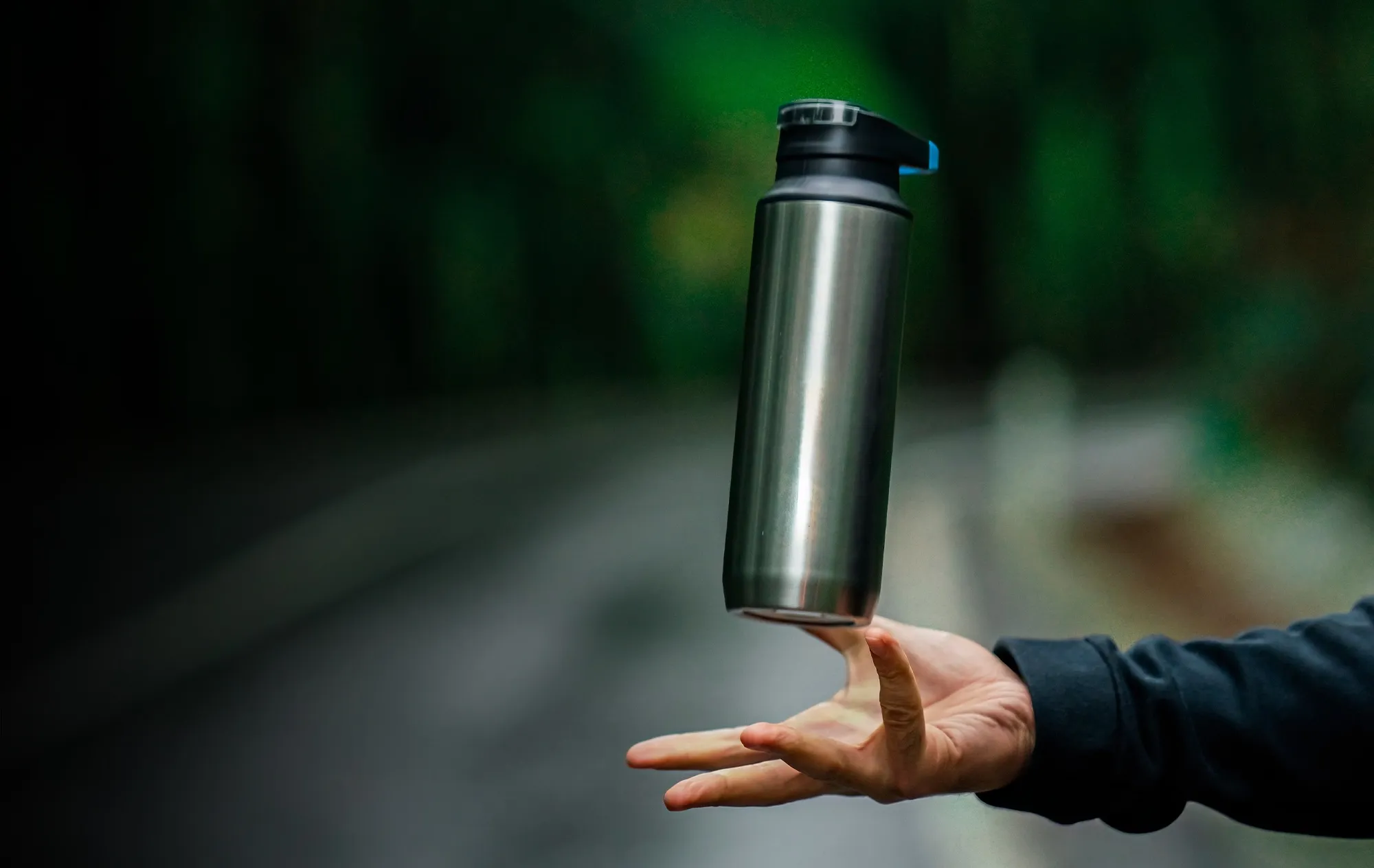 A stainless steel drink bottle with a black lid hovering just above an open hand, against blurred green trees