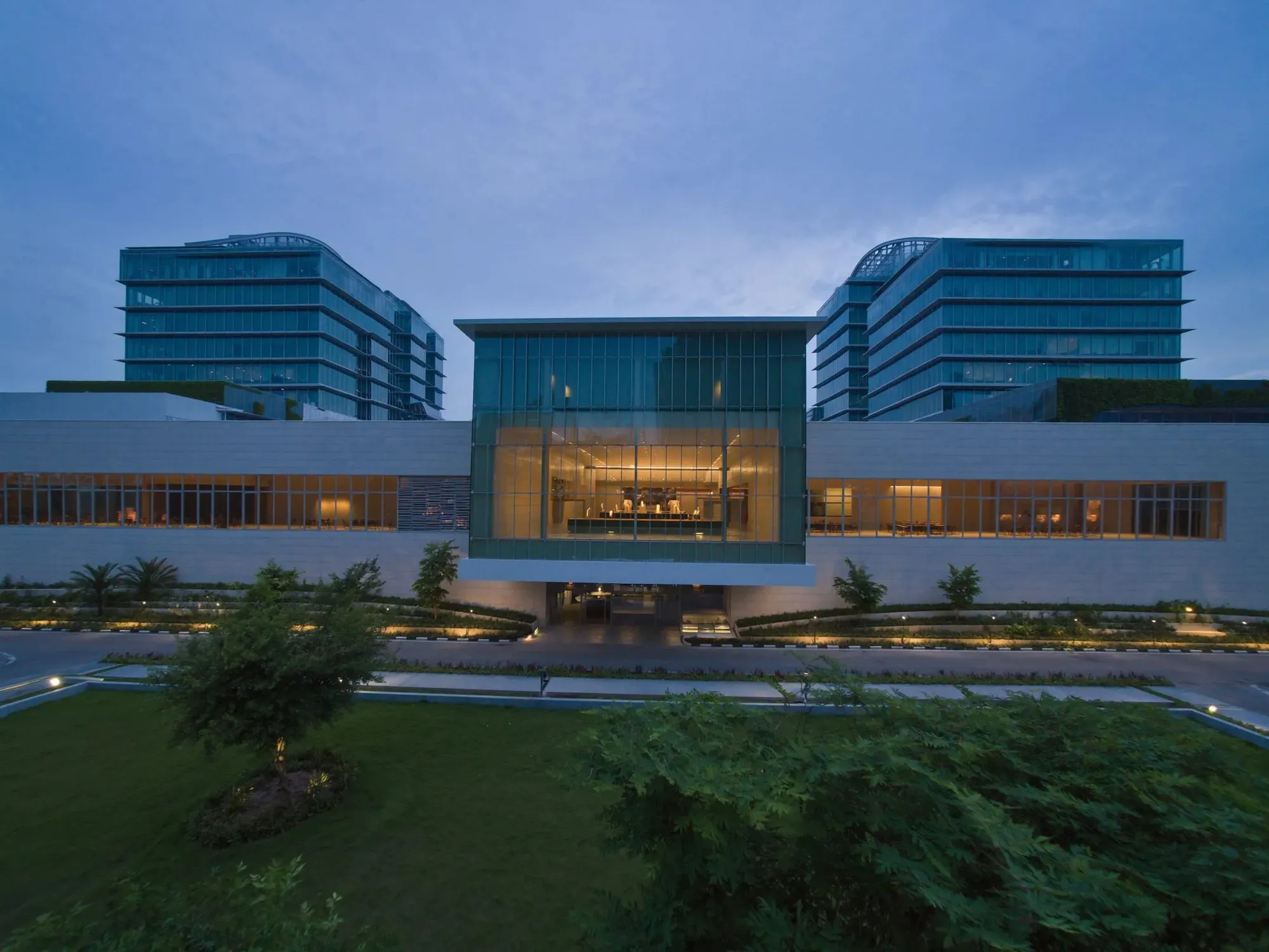 The front of a modern hospital at dusk, a lit glass entrance hall between two glass towers, trees and a lawn in front